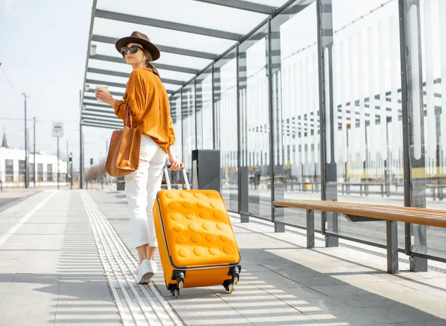 Young female traveler walking with a yellow suitcase at the modern transport stop outdoors, back view. Concept of an urban transportation and travel