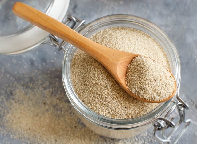 Raw uncooked fonio seeds in a glass jar with a spoon on grey background close up