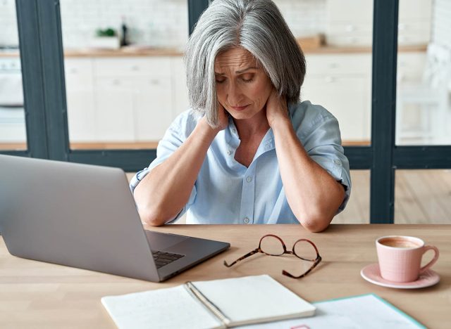 Tired stressed old mature business woman suffering from neckpain working from home office sitting at table. Overworked senior middle aged lady massaging neck feeling hurt pain from incorrect posture.