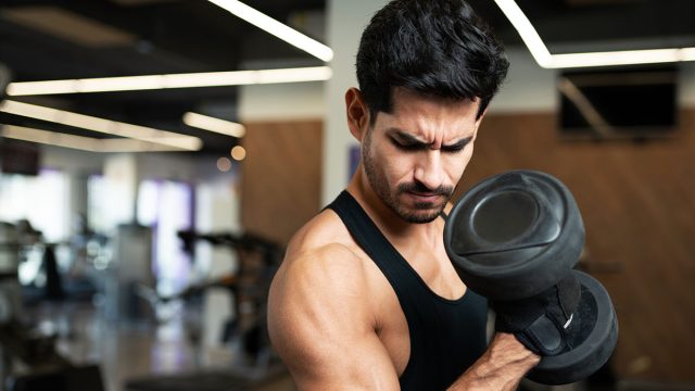 Hispanic young man looking at his arm muscle while doing a bicep curl with weights at the fitness center