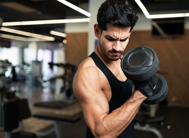 Hispanic young man looking at his arm muscle while doing a bicep curl with weights at the fitness center