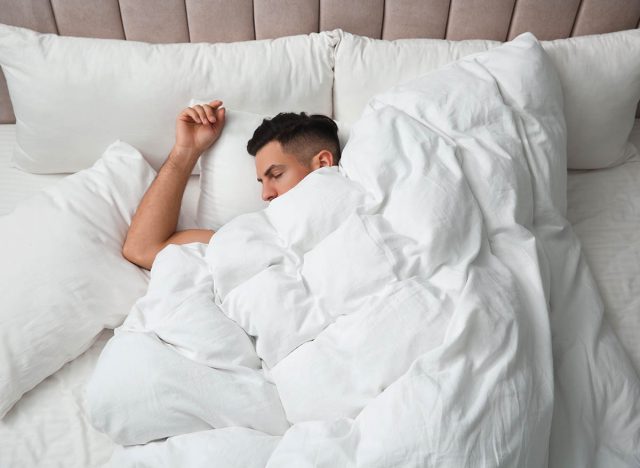 Handsome man sleeping under soft blanket in bed at home, above view