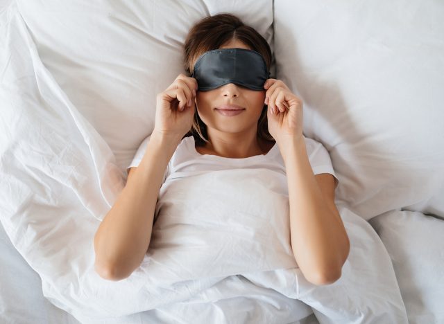 Pleasant young woman laying in bed and putting eye mask on before going to sleep