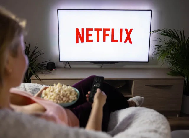 Woman holding popcorn and watching TV at home with Netflix logo on the screen.