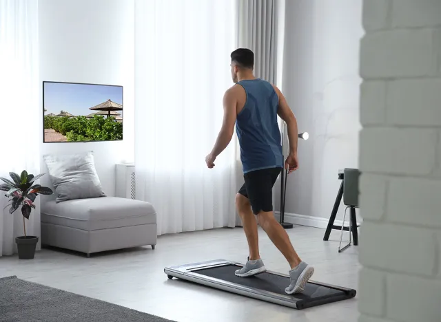 Sporty man training on walking treadmill and watching TV at home