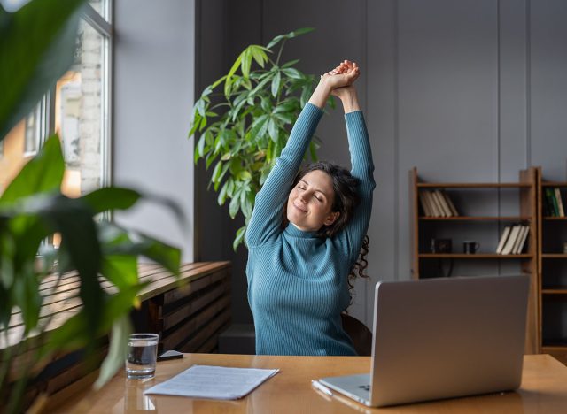 Happy businesswoman warming up body and muscles at workplace, feeling satisfied with work done, smiling female employee resting from computer screen. Well-being, productivity and happiness at work