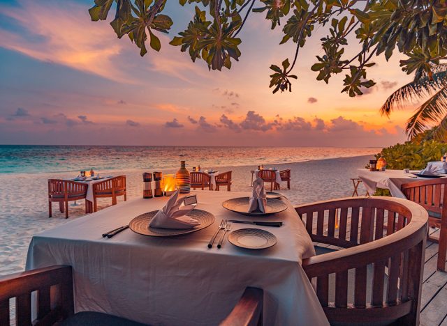 Outdoor restaurant at the beach. Table setting at tropical beach restaurant. Led light candles and wooden tables, chairs under beautiful sunset sky, sea view. Luxury hotel or resort restaurant