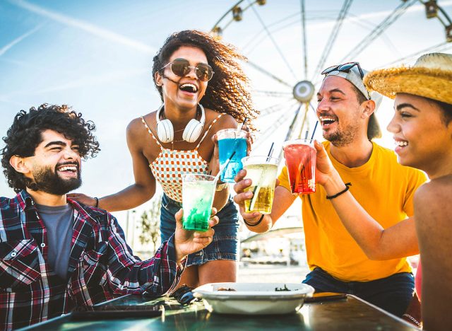 Happy young people cheering cocktail glasses together at beach party - Multi-ethnic friends enjoying happy hour sitting at bar table - Youth lifestyle and summertime vacations concept - Focus on eyes