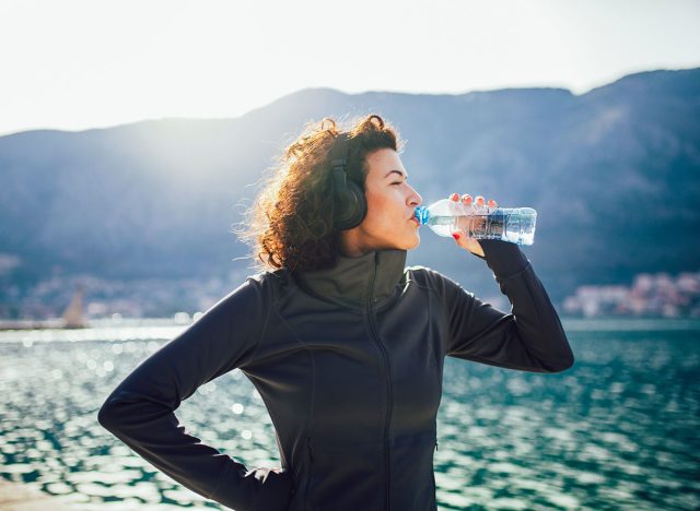 Fitness woman drinking water from bottle. Muscular young female taking a break from workout outside.