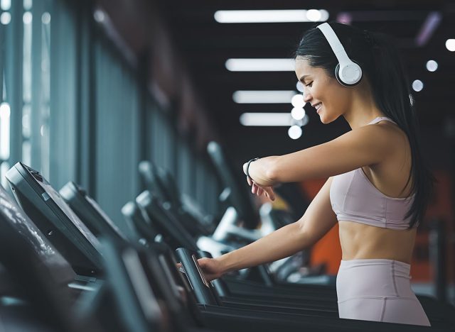 Panoramic Side view of beautiful young asian woman running on treadmill and listening to music via headphone with arm a smart watch for tracking speed during sports training in a gym.