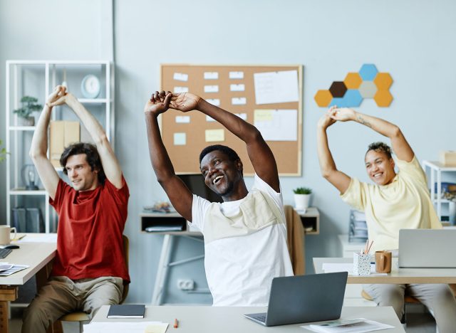 Diverse group of smiling young people doing stretching exercises at workplace in office