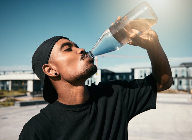 City heat, summer and black man drinking water on concrete road, thirsty gen z outdoor activity. Sun, fun and urban young man with healthy street culture lifestyle and clean water in bottle to drink.