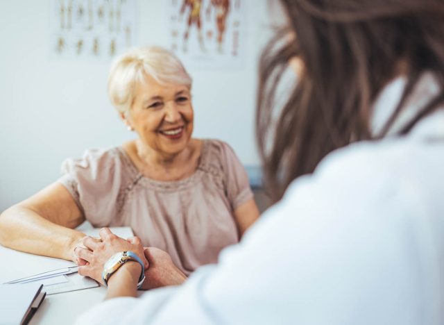 Doctor giving hope. Close up shot of young female physician leaning forward to smiling elderly lady patient holding her hand in palms. Woman caretaker in white coat supporting encouraging old person