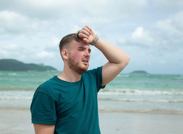 Guy is suffering from heat, man with heatstroke. Having sunstroke at summer hot weather. Dangerous sun, boy under sunshine. Headache, feeling bad. Person holds hand on head at sea, beach