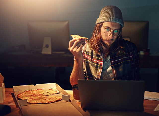 Web designer, man with pizza at laptop and night shift, overtime and deadline at digital marketing agency. Content research, reading and serious male at desk, working late at startup eating fast food