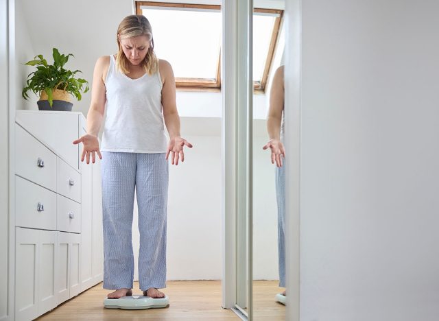 Menopausal Mature Woman Concerned With Weight Gain Standing On Scales In Bedroom At Home