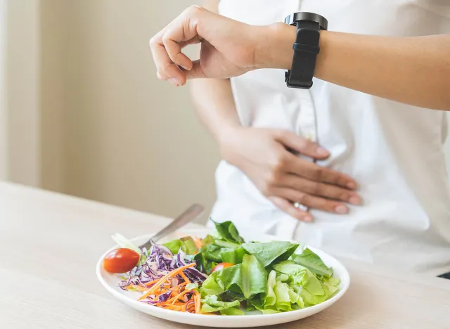 Intermittent fasting, health care asian young woman dietary, having stomach ache, temptation hungry of breakfast food in morning on table at home, looking at watch on her wrist but not yet time to eat