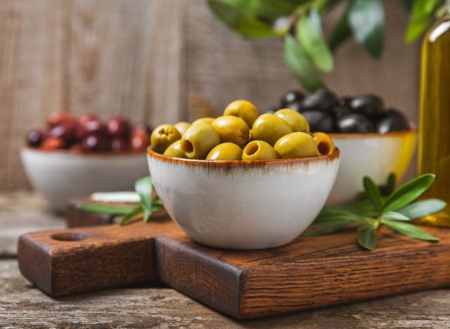 Green, black and red olives, olive oil on a brown wooden background. Fresh juicy olives in a bowl and fresh olive leaves. Vegan. Olive fruits. Place for text. Copy space.