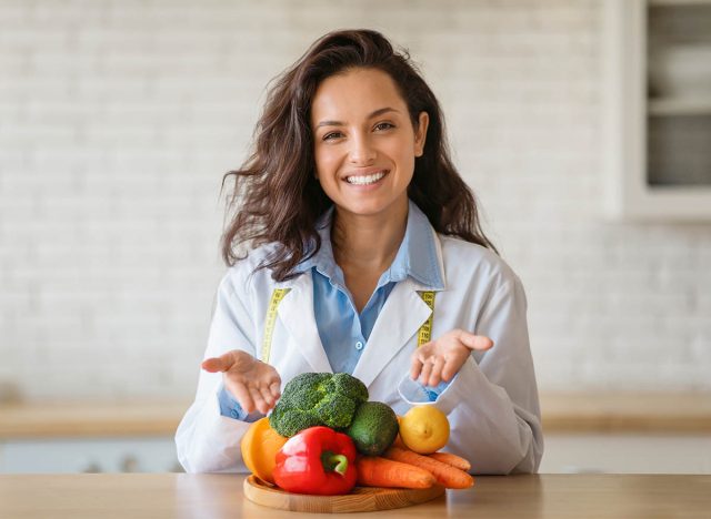 Portrait of cheerful dietitian in lab coat pointing at fresh fruits and vegetables on table, smiling at camera at clinic. Weight loss consultant recommending healthy plant based diet