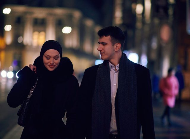 Happy multicultural business couple walking together outdoors in an urban city street at night near a jewelry shopping store window.