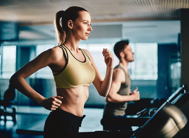 Pretty woman running on treadmill with fit young man on background