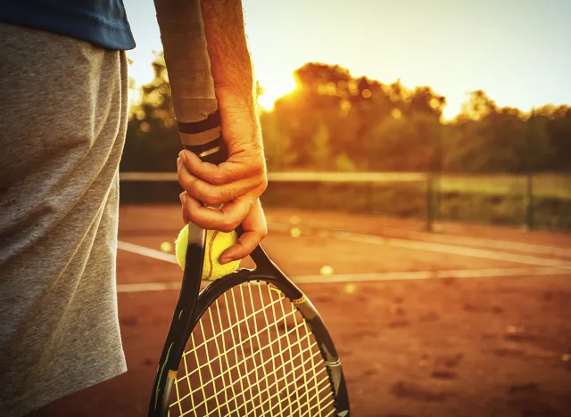 Close up of man holding tennis racket on clay court. In his hand is tennis ball. On court is sunset./Man holding tennis racket