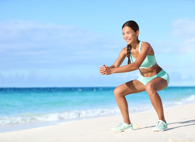 Fitness young woman working out core and glutes with bodyweight workout doing squat exercises on beach. Asian sporty girl squatting legs as part of an active and fit life.