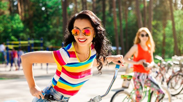 Outdoor lifestyle portrait of two best friends hipster girls wearing stylish bright outfits, denim shorts and glasses going crazy and having great time together. Laughing and ride bikes
