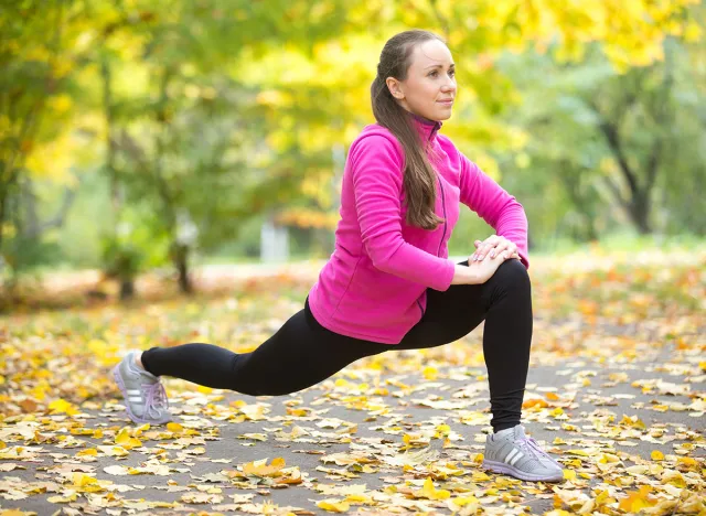 Sporty beautiful happy young woman warming up, stretching before morning running routine, doing high lunges, exercises for legs, hips and buttocks, working out outdoor on autumn day. Full length