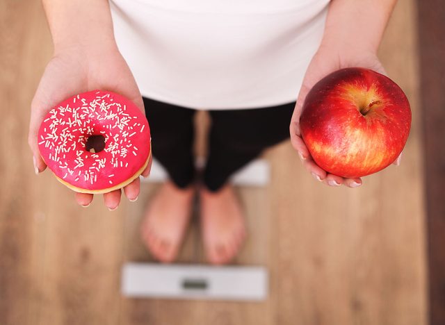 Diet. Woman Measuring Body Weight On Weighing Scale Holding Donut and apple. Sweets Are Unhealthy Junk Food. Dieting, Healthy Eating, Lifestyle. Weight Loss. Obesity. Top View