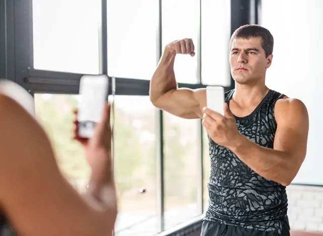 Portrait of proud bodybuilder boasting his arm muscles taking selfie in gym mirror flexing biceps after working out