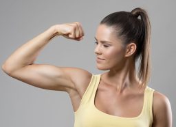 Strong sporty fit woman in yellow tank top flexing bicep muscle over gray studio background.