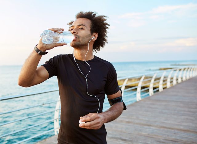 Stylish Afro-American male runner drinking water out of plastic bottle after cardio workout, wearing white earphones. Sportsman in black sportswear hydrating during outdoor training.
