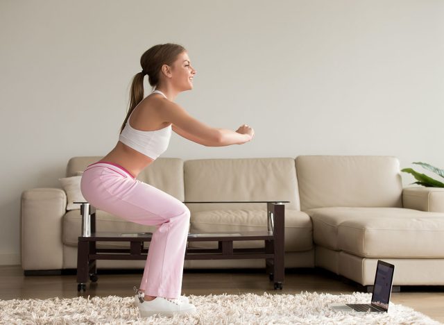 Young sporty woman doing two-leg deep squat morning exercise standing in living room, teen girl burning fat calories with online fitness training help crouching for buttocks lift at home, side view