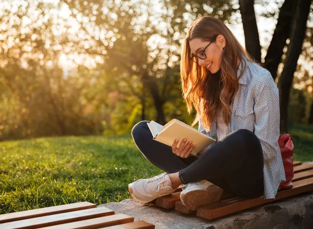 Side view of pleased brunette woman in eyeglasses sitting on bench and reading book in park