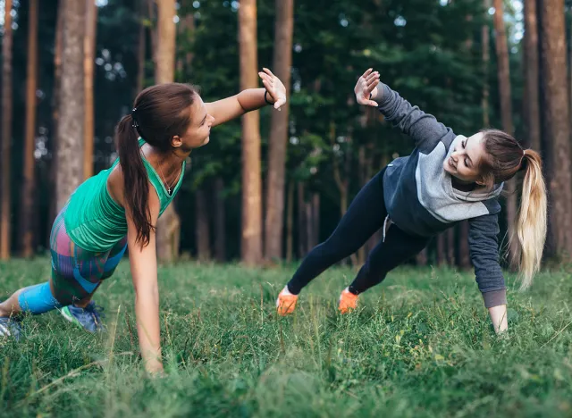 Two female buddies doing partner side plank giving high five while training in the forest
