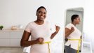 Plus size black woman measuring waist with tape, showing results of slimming diet, standing in front of mirror, gesturing thumb up at home. Young lady promoting healthy nutrition for weight loss