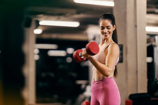 A muscular sportswoman is doing dumbbells curls in a gym and flexing muscles.