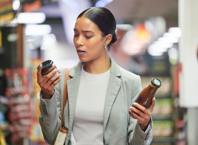 Woman in a store or supermarket, reading product labels of choice to decide or compare sauce bottles.
