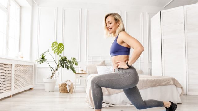Athletic woman in stylish sportswear doing lunges exercises at home in bedroom.