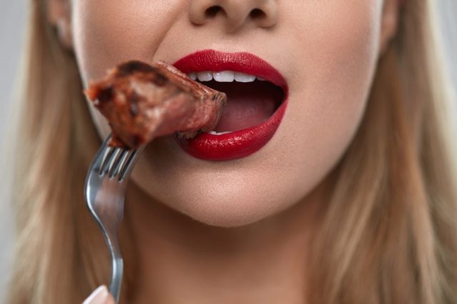 Eating Meat. Closeup Of Woman Mouth With Red Lips, White Teeth Biting Tasty Beef Steak On Fork. Close-up Of Beautiful Female Mouth Eating Delicious Grilled Meat. Nutrition Concept. High Resolution