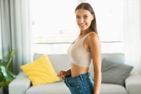 Young slim woman enjoying successful weight loss, showing great result wearing old large jeans for comparison, smiling to camera.