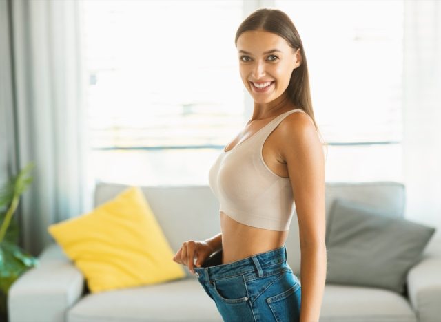 Young slim woman enjoying successful weight loss, showing great result wearing old large jeans for comparison, smiling to camera.