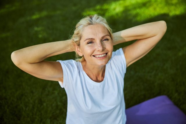 Mature lady in white shirt holding hands behind her head and expressing positive emotions while spending time outdoors.