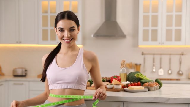 Happy woman measuring waist with tape in kitchen.