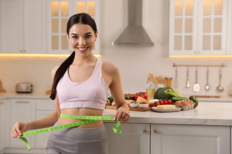 Happy woman measuring waist with tape in kitchen.