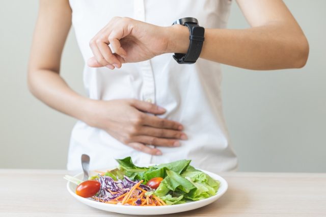 Intermittent fasting, health care asian young woman dietary, having stomach ache, temptation hungry of breakfast food in morning on table at home, looking at watch on her wrist but not yet time to eat