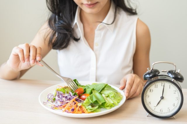 Intermittent fasting with clock, health asian young woman, girl weight loss, eating green fresh vegetable salad on dish, plate with eat healthy of breakfast food in morning, lunch on a table at home.