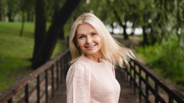 Smiling cheerful beautiful blonde caucasian mature woman looking at the camera while walking in city public forest wood park.