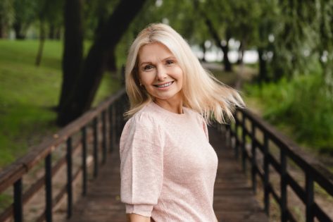 Smiling cheerful beautiful blonde caucasian mature woman looking at the camera while walking in city public forest wood park.